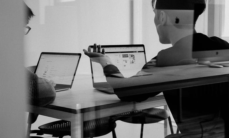 Two people sitting at a table talking in front of their laptops